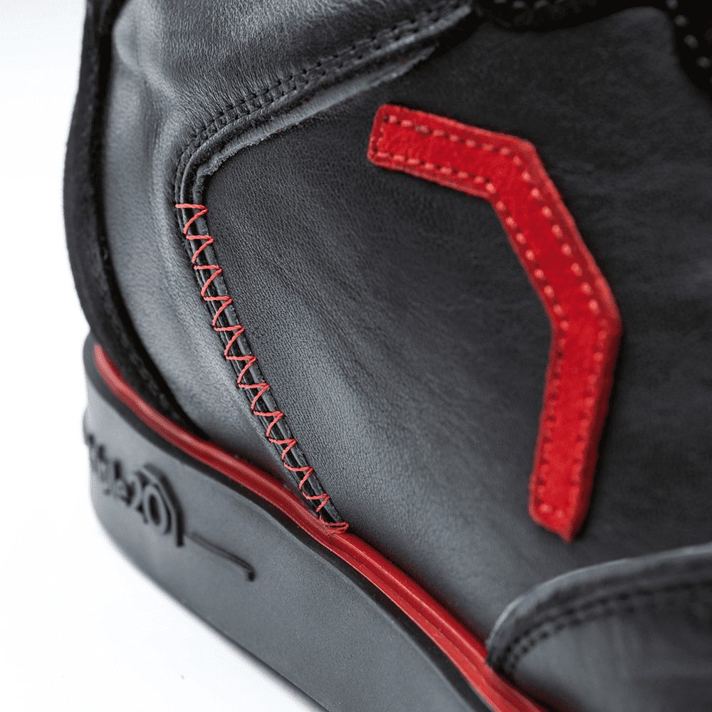 Der Schuh besteht aus schwarzem Leder mit auffälligen roten Nähten und Applikationen. Auf der Sohle ist zudem ein roter Streifen zu sehen.