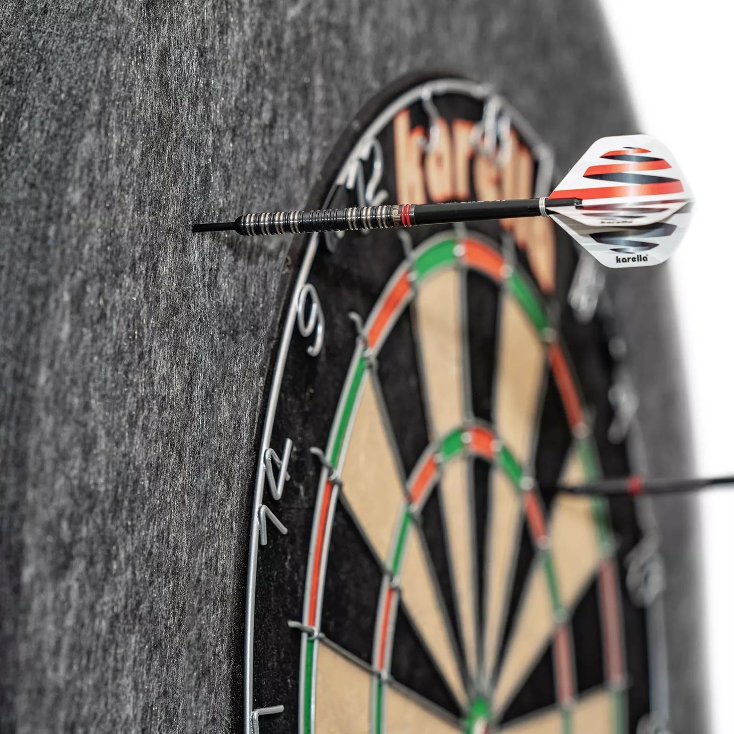 Karella Ultimo steel dartboard surround Das Bild zeigt eine Karella Ultimo Steeldartboard Surround, die einen Dartpfeil auffängt. Die graue Umrandung schützt die Wand vor Fehltreffern neben der Dartscheibe.