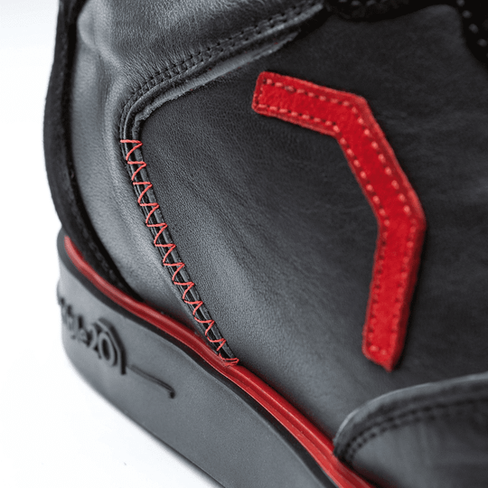 Der Schuh besteht aus schwarzem Leder mit auffälligen roten Nähten und Applikationen. Auf der Sohle ist zudem ein roter Streifen zu sehen.